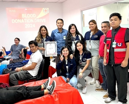 A group of 10 men and women smiling at the camera in a building with a sign on the wall that reads 'Blood Donation Drive 2024.' To the right are four people laying down on red cots
