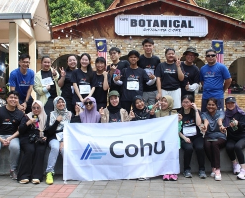 A group of 21 men and women smiling at the camera holding up peach signs and some holding up a white flag that reads 'Cohu.' They are outside and in the background is a building with Malaysia flags and a sign reading 'Kafe Botanical Gateway Cafe'