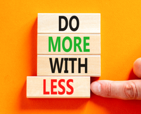 An orange background with four wooden blocks arranged vertically. Each one reads one word 'Do' 'More' 'With' 'Less' and there is a hand pushing the last block 'Less' out of alignment with the others
