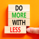 An orange background with four wooden blocks arranged vertically. Each one reads one word 'Do' 'More' 'With' 'Less' and there is a hand pushing the last block 'Less' out of alignment with the others