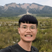 A headshot of a man wearing glasses and smiling with a mountain and grassy landscape in the background