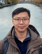 A man looking at the camera wearing glasses and layers including a dark brown winter coat with a body of water in the background