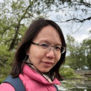 Headshot of a woman wearing glasses and a pink puffer jacket with trees and a river in the background